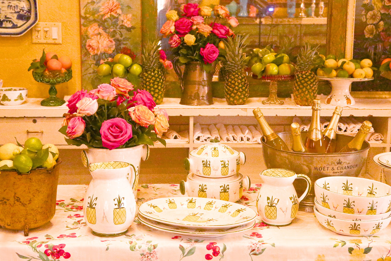 The Ivy Pineapple pattern tableware with hand-painted yellow pineapples on ceramic bowls, pitchers, and plates, styled with roses, citrus, and champagne.