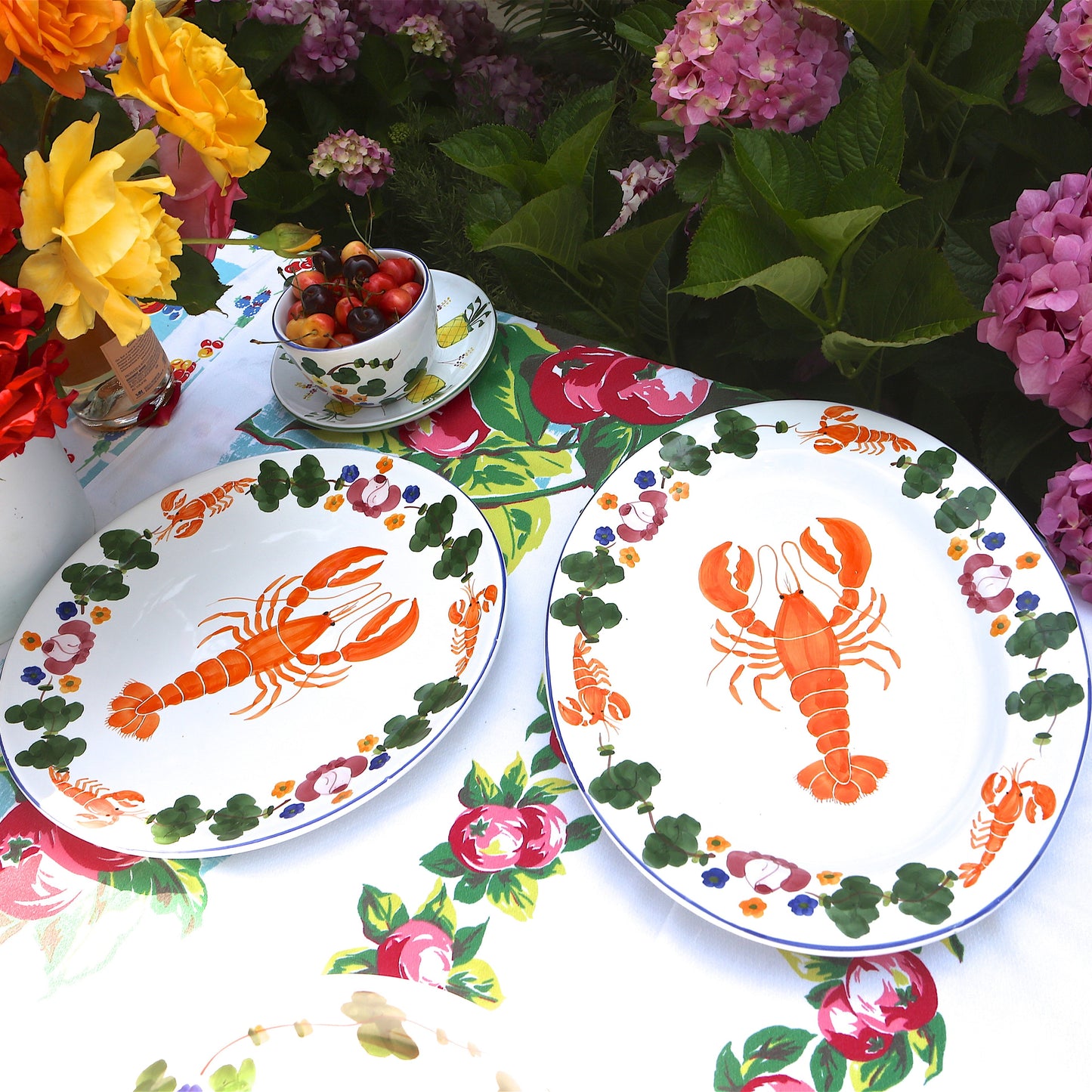 Two plates with lobster design on a tablecloth with floral patterns and flowers in the background.