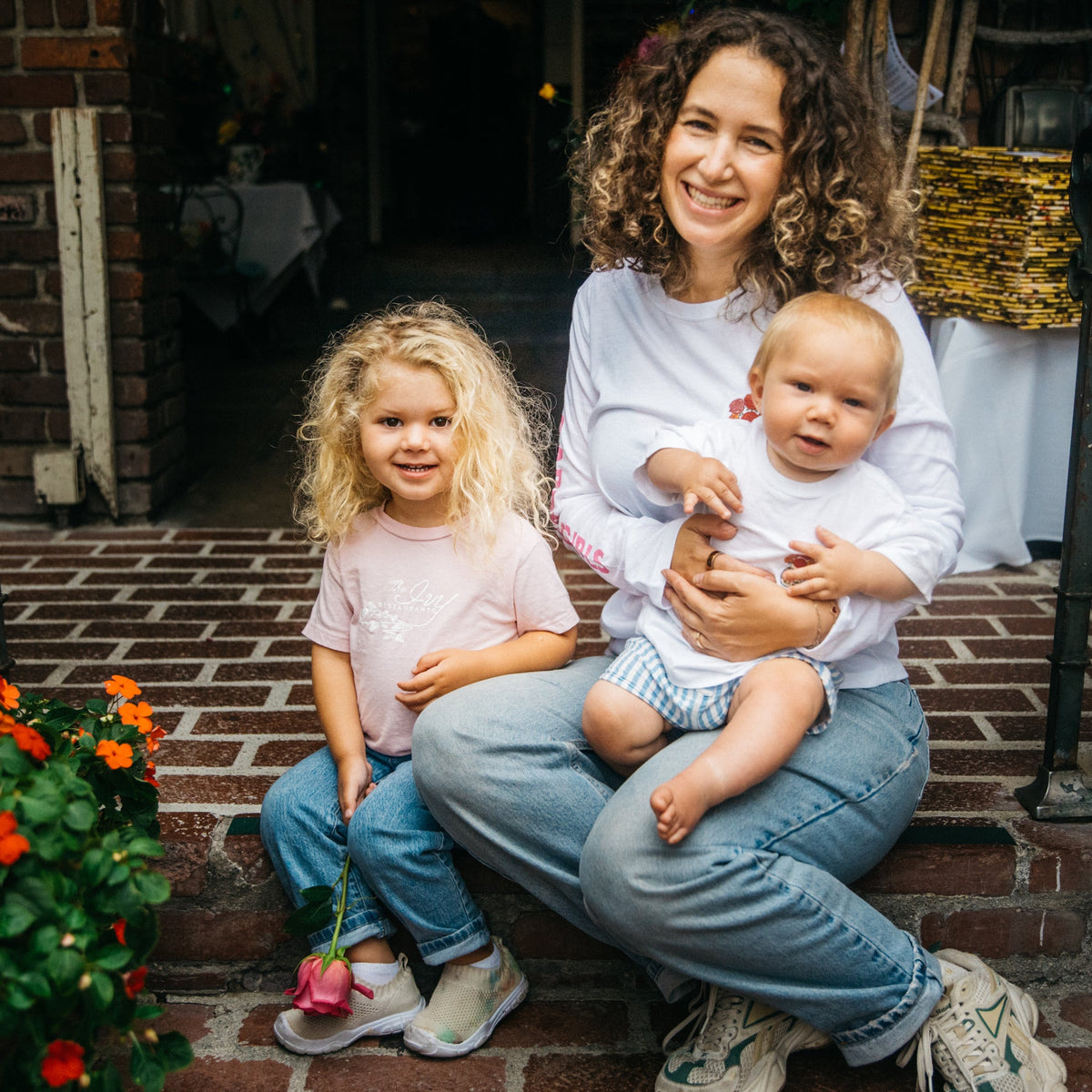 A smiling woman with curly hair sits on brick steps holding a baby, with a young girl standing beside them. The woman wears a white long-sleeve top and light blue jeans, while the girl wears a light pink top and blue jeans.
