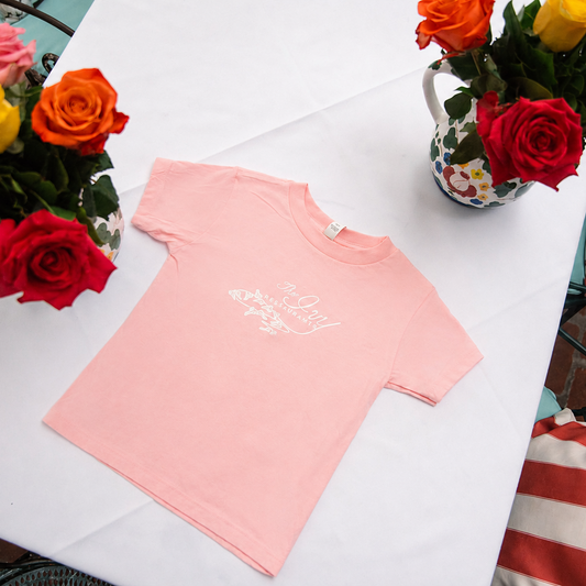 A light pink short-sleeve t-shirt with white cursive text on the chest, laid flat on a white tablecloth. The shirt is styled with colorful rose bouquets in vases and patterned pillows in the background.
