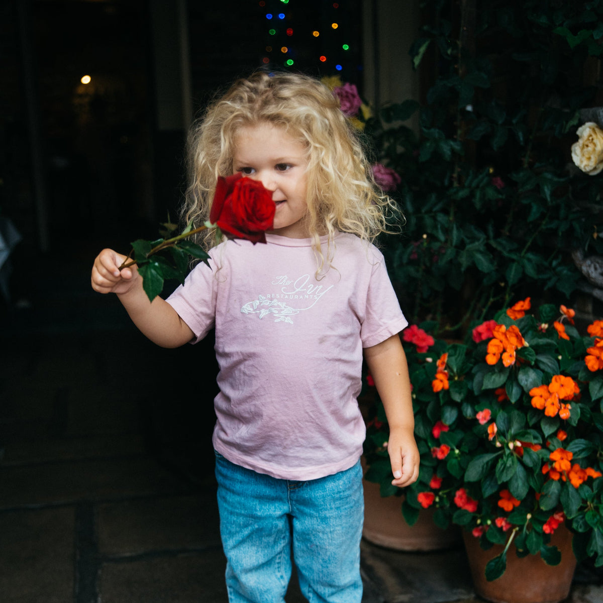 A young child with curly blonde hair stands on a brick porch holding a red rose near their face. The child is wearing a light pink t-shirt, blue jeans, and white socks, with potted flowers and a dark doorway in the background.