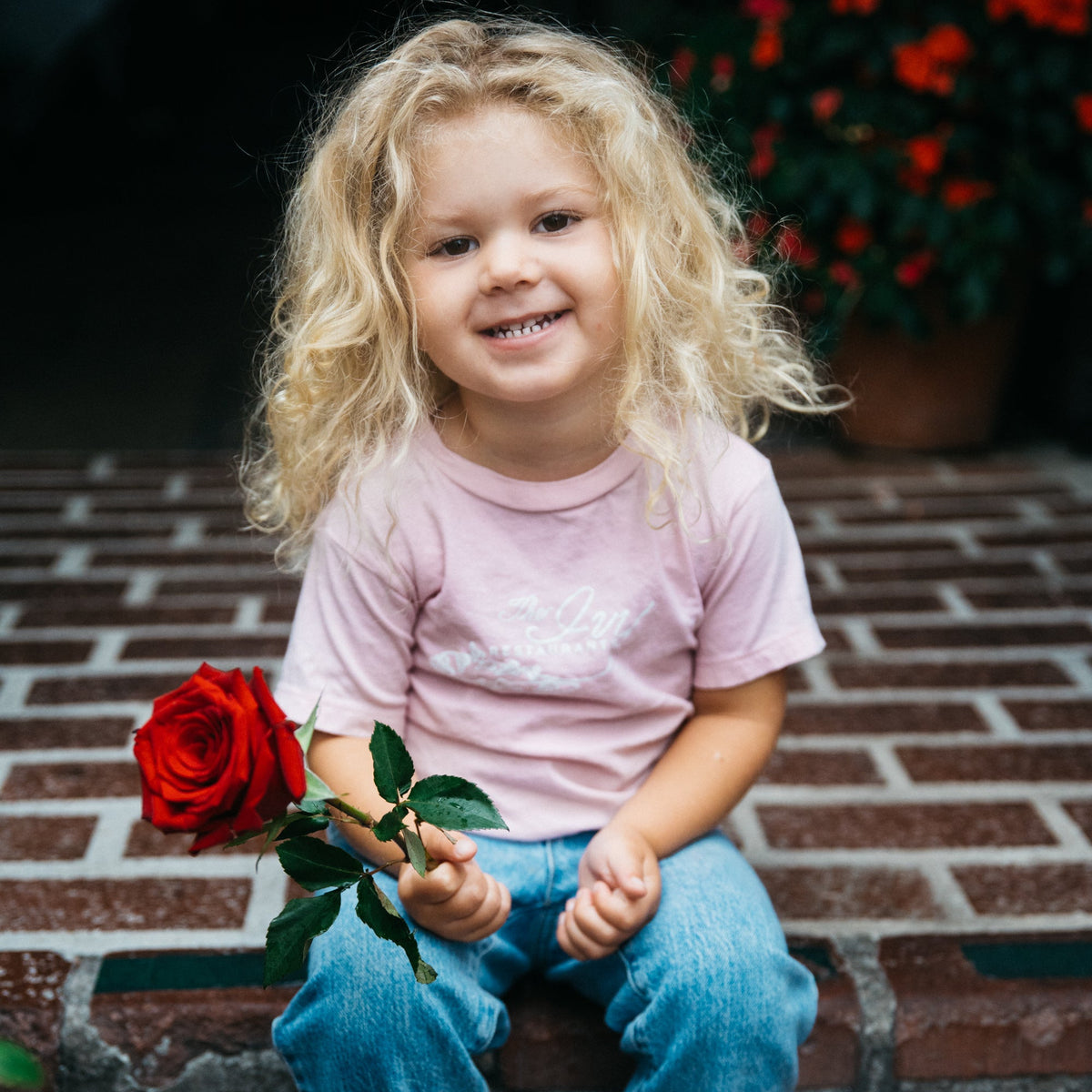 A young girl with curly blonde hair sits on red brick steps holding a single red rose. She is wearing a light pink t-shirt, and blue jeans.