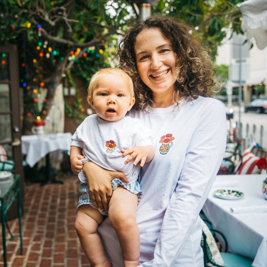 A smiling woman with curly hair holds a baby outdoors on a patio. Both are wearing matching white long-sleeve shirts with a colorful floral chest graphic.