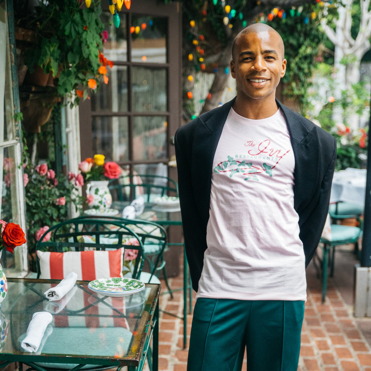A smiling man stands on a brick patio at a restaurant, wearing a white graphic t-shirt under a black blazer and dark green trousers.