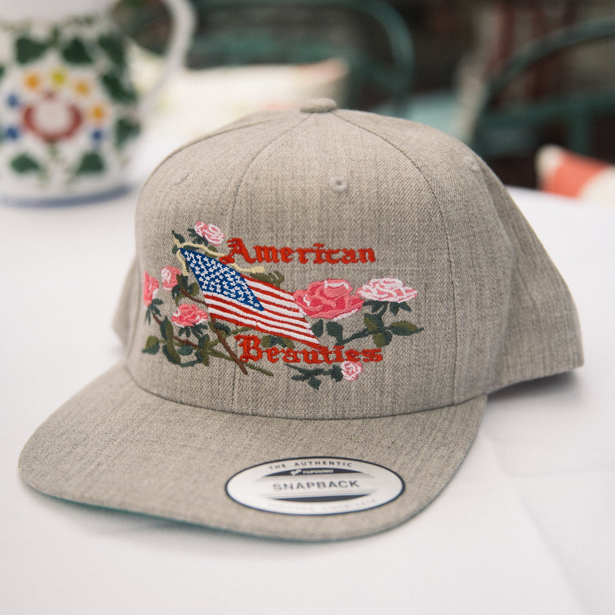 A light gray heather snapback cap with an embroidered design featuring a red and white American flag, pink roses, green leaves, and the words 'American Beauty' in red script. The cap is displayed on a white surface with a blurred background containing a floral mug and green chairs.