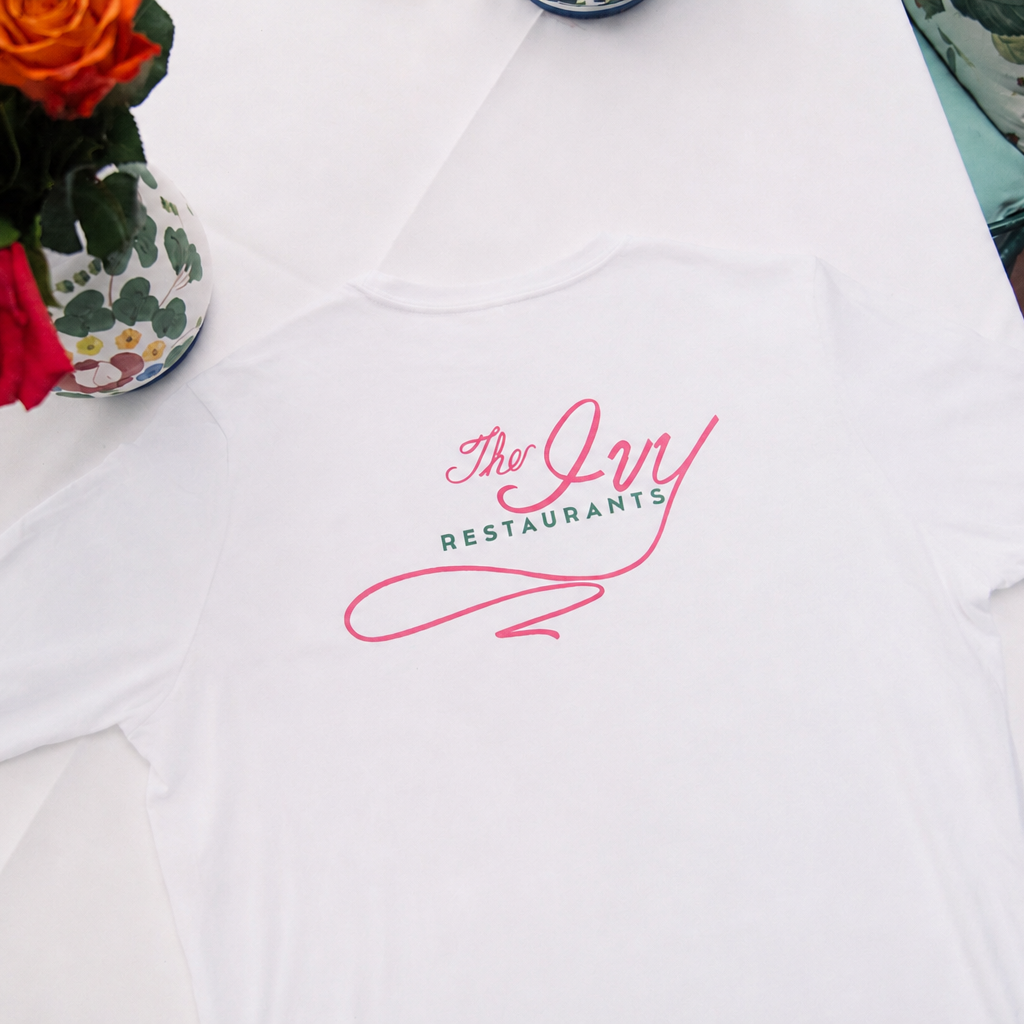A white short-sleeve T-shirt laid flat on a table, featuring a pink and green printed logo that reads 'The Ivy Restaurants' on the chest. Decorative red and orange roses in a patterned vase sit in the upper left corner.