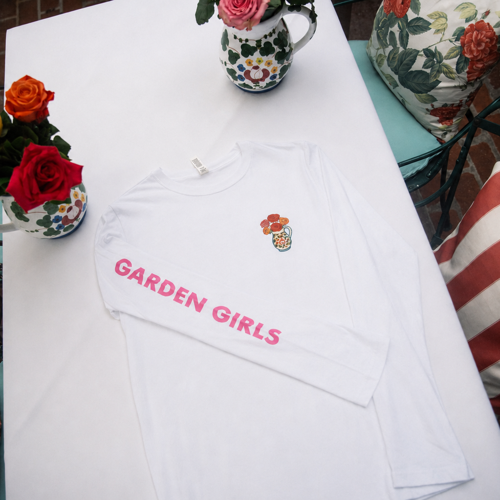 A white long-sleeve t-shirt with 'GARDEN GIRLS' printed in pink across the chest and a small colorful floral graphic on the left chest, laid flat on a white table with decorative flowers and a floral pillow in the background.